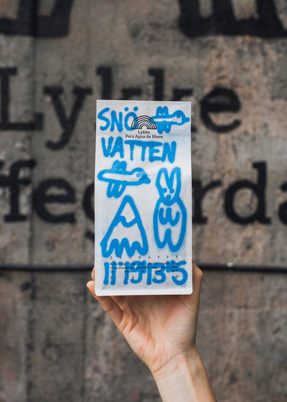 Someone holding a white coffee bag with blue graffiti-style art and writing on, including "Snövatten", against a grey, industrial background.